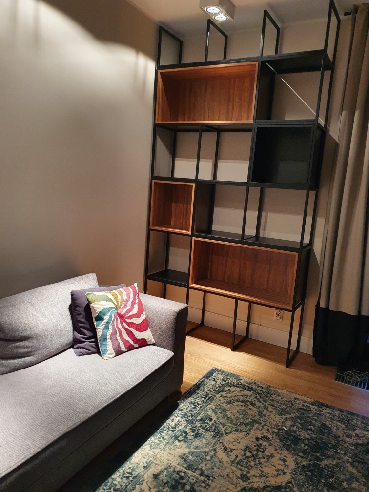 Regał metalowy z drewnianymi wstawkami, stojący obok szarej sofy z dekoracyjnymi poduszkami, na tle wzorzystego dywanu.