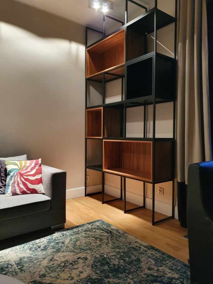 Regał modułowy z czarnej stali i drewnianymi półkami, widoczny w salonie z szarą sofą i wzorzystym dywanem.