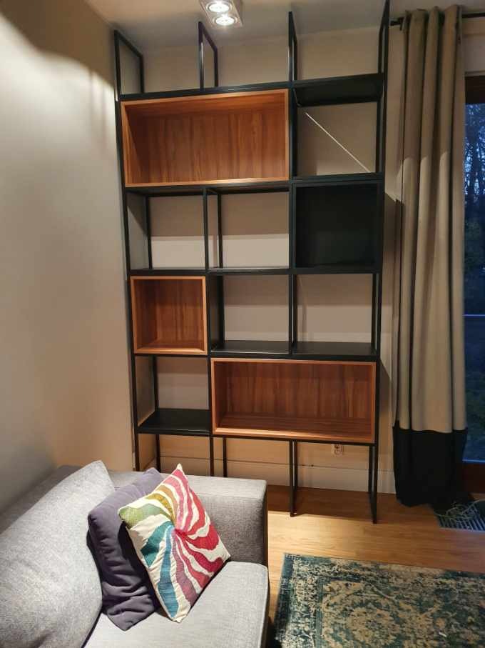 Ręcznie wykonany regał z czarnego metalu i drewnianych półek, ustawiony w salonie obok szarej sofy z kolorową poduszką.