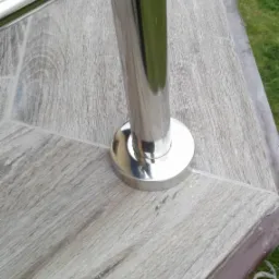 Szczegół mocowania słupka ze stali nierdzewnej do podłoża imitującego drewno, widoczny fragment trawnika w tle.