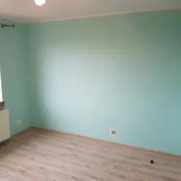 Pomalowane na miętowo ściany w pustym pokoju z panelami podłogowymi, widoczny grzejnik pod oknem i gniazdko elektryczne na ścianie.