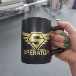 Czarny kubek z nadrukiem złotego logo i napisu 'OPERATOR' trzymany w dłoni z zielonymi paznokciami, w tle maszyny drukarskie.