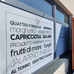 Grafika okienna z menu pizzerii wypisanym w różnych krojach pisma, naklejona na witrynę lokalu, widoczna z zewnątrz w słoneczny dzień.