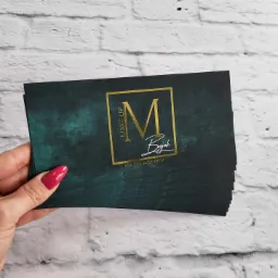Stos wizytówek z logo studia makijażu w ręce z czerwonym manicure na tle białej cegły.