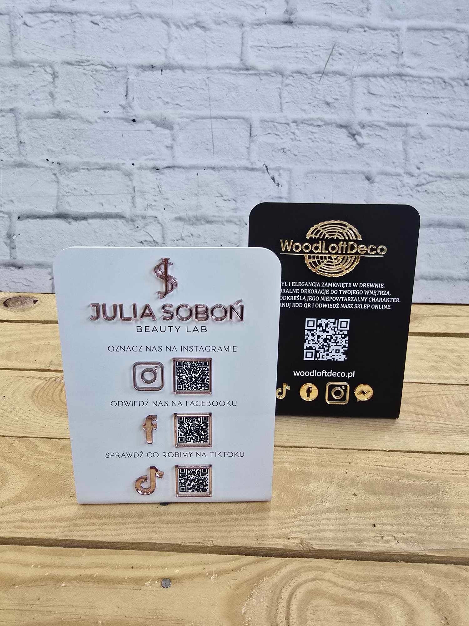 Dwa stojaki reklamowe: biały z logo Julia Soboń Beauty Lab i czarny z logo WoodLoftDeco, prezentujące kody QR do mediów społecznościowych na drewnianym blacie.
