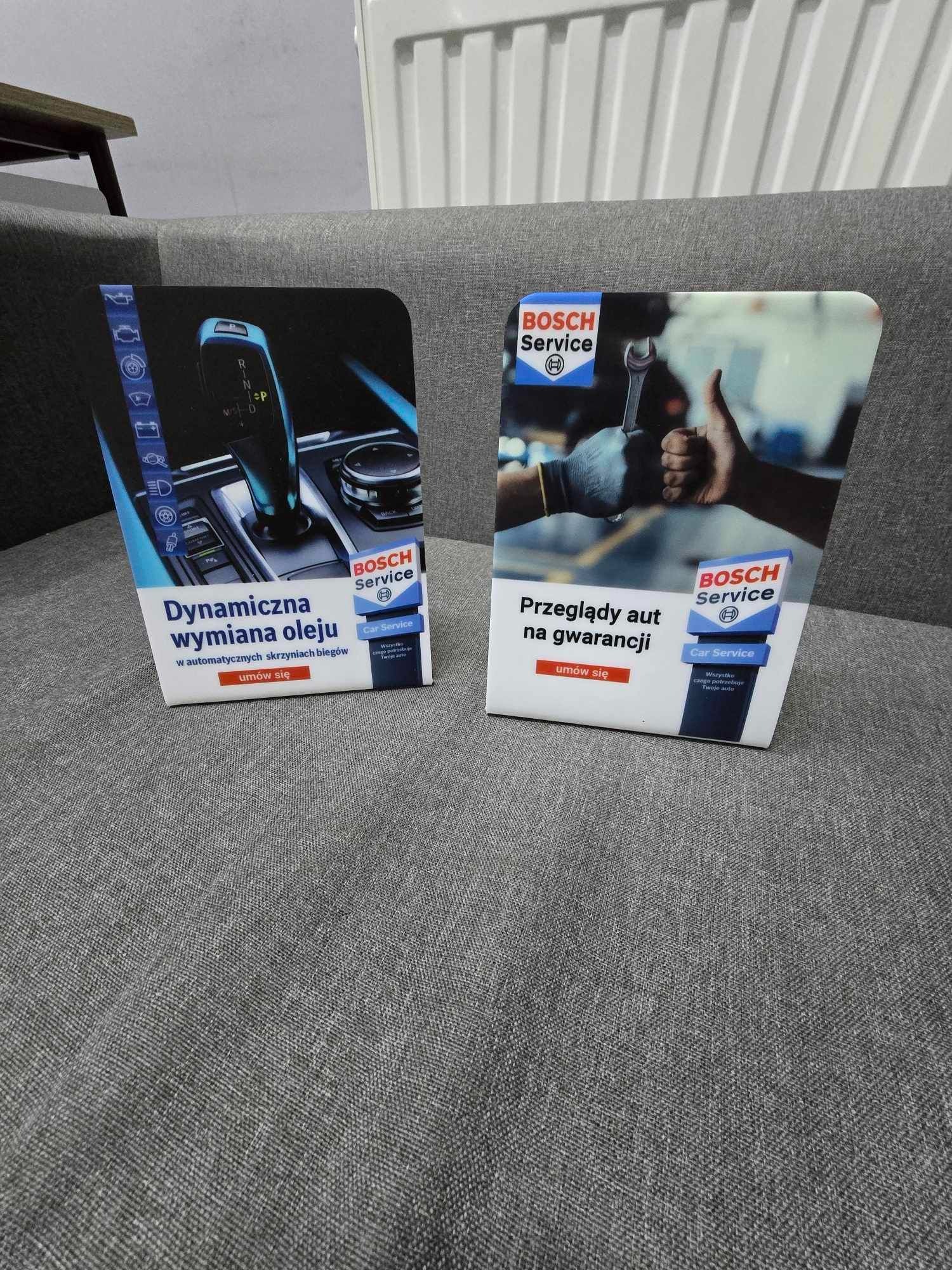 Dwa stojące, laminowane panele reklamowe Bosch Service na szarym tle sofy. Jeden panel promuje dynamiczną wymianę oleju w automatycznych skrzyniach biegów, drugi przeglądy aut na gwarancji.