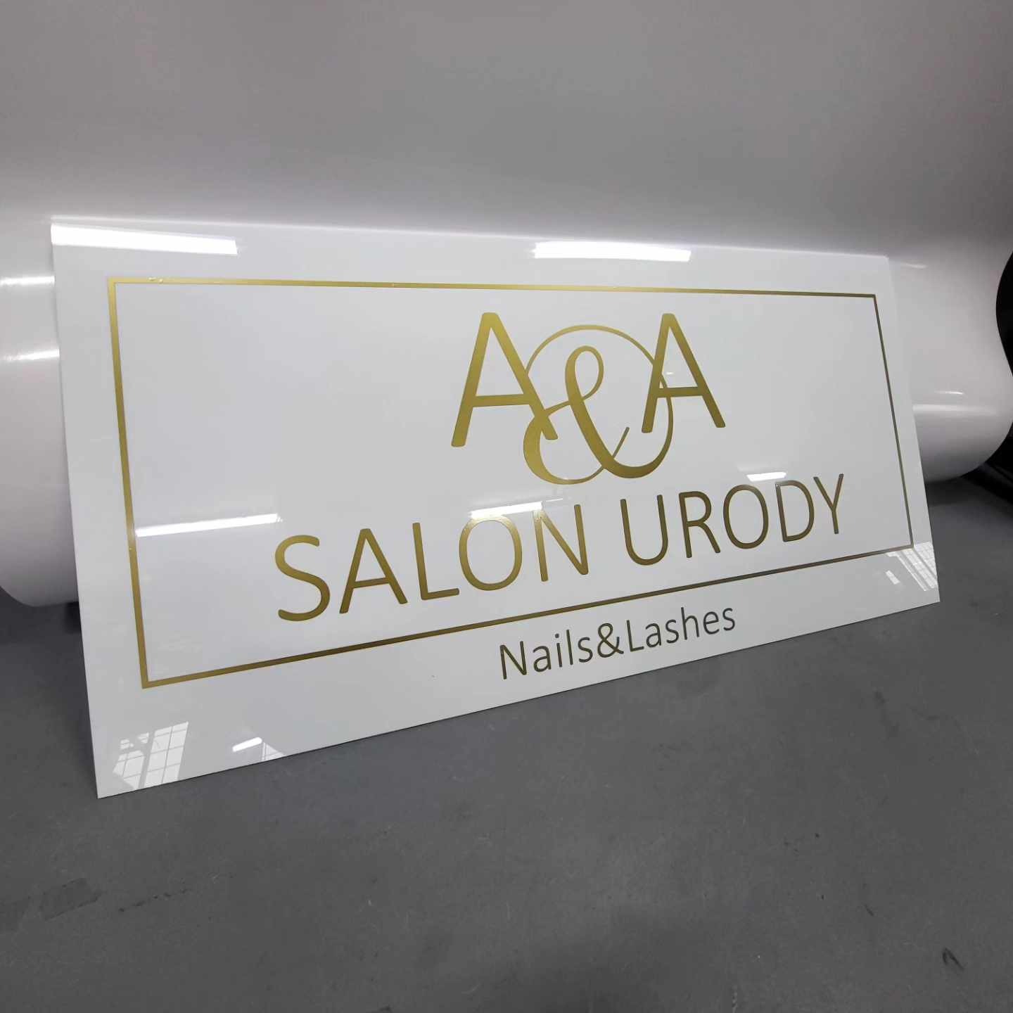 Biała tablica informacyjna z złotym napisem 'A&A SALON URODY Nails&Lashes' w prostokątnej złotej ramce, umieszczona na szarym tle, lekki kąt ujęcia.