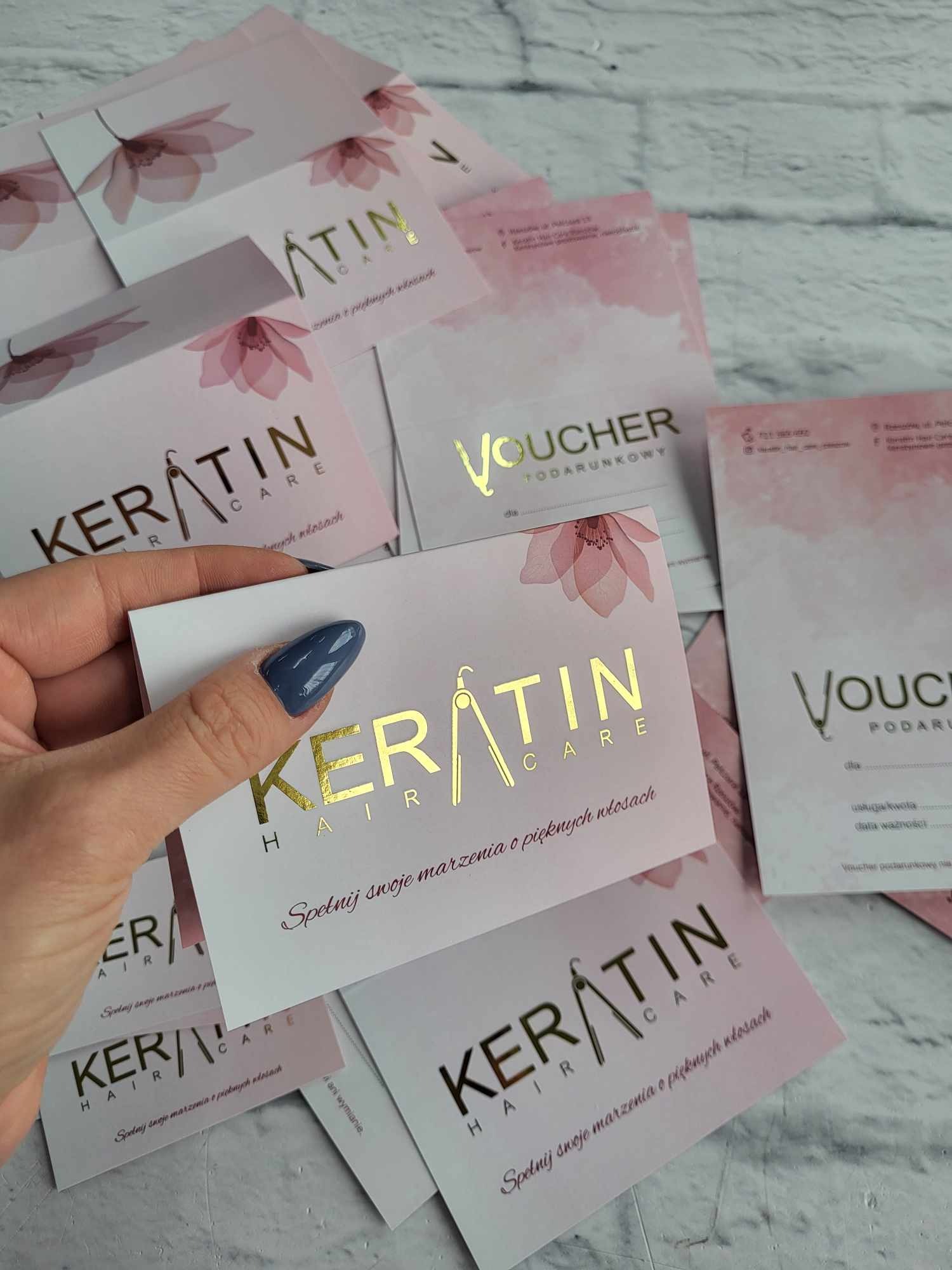 Stos wizytówek i voucherów salonu fryzjerskiego Keratin Hair Care w odcieniach różu i złota, trzymanych w dłoni z niebieskim lakierem do paznokci.