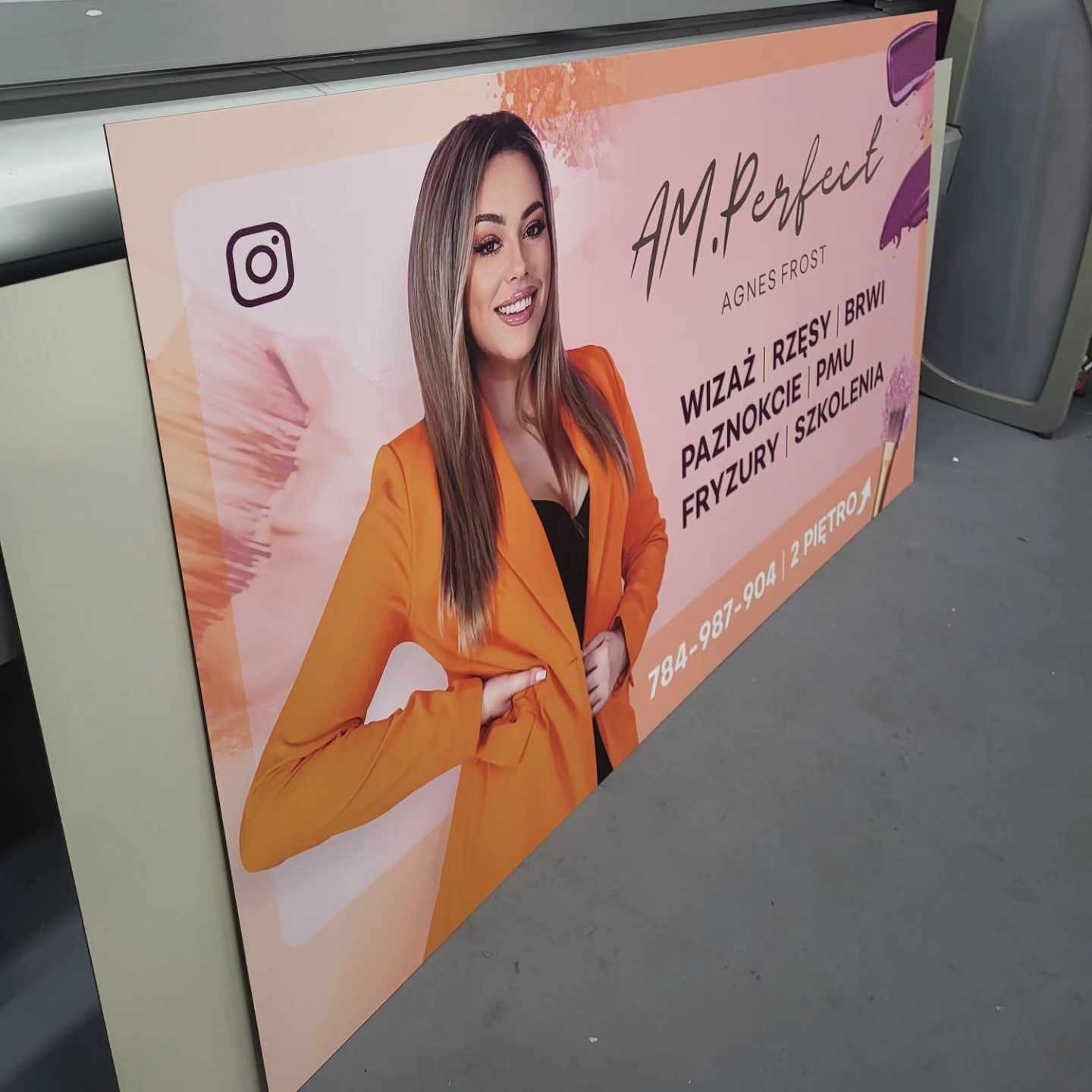 Wydrukowany baner reklamowy z wizerunkiem kobiety w pomarańczowym żakiecie, prezentujący usługi wizażu, stylizacji rzęs, brwi, paznokci, makijażu permanentnego, fryzur i szkoleń.