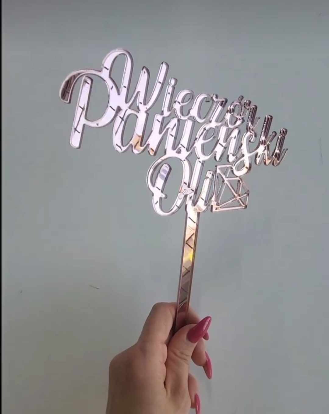 Ręka trzymająca złoty topper na tort z napisem 'Wieczór Panieński Oli' z motywem diamentu, na tle jasnej ściany.