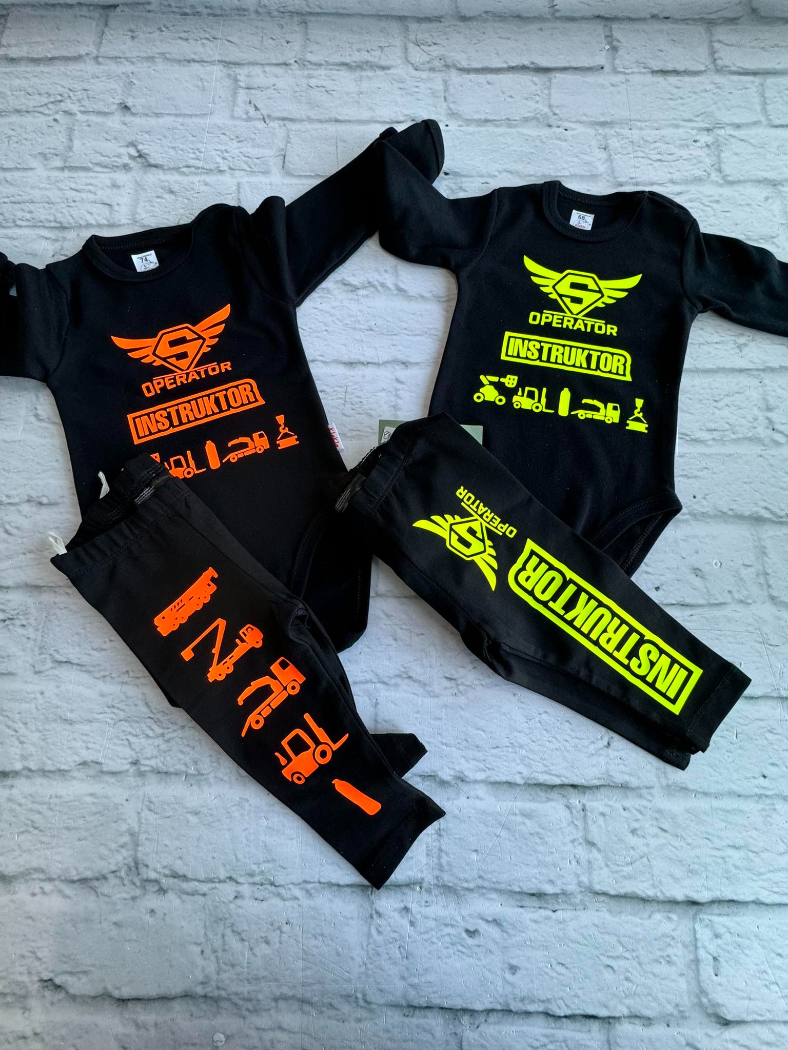 Dwa czarne niemowlęce body z długim rękawem i legginsy z motywem 'Operator Instruktor' w kolorze pomarańczowym i neonowej zieleni, leżące na tle ściany z białej cegły.