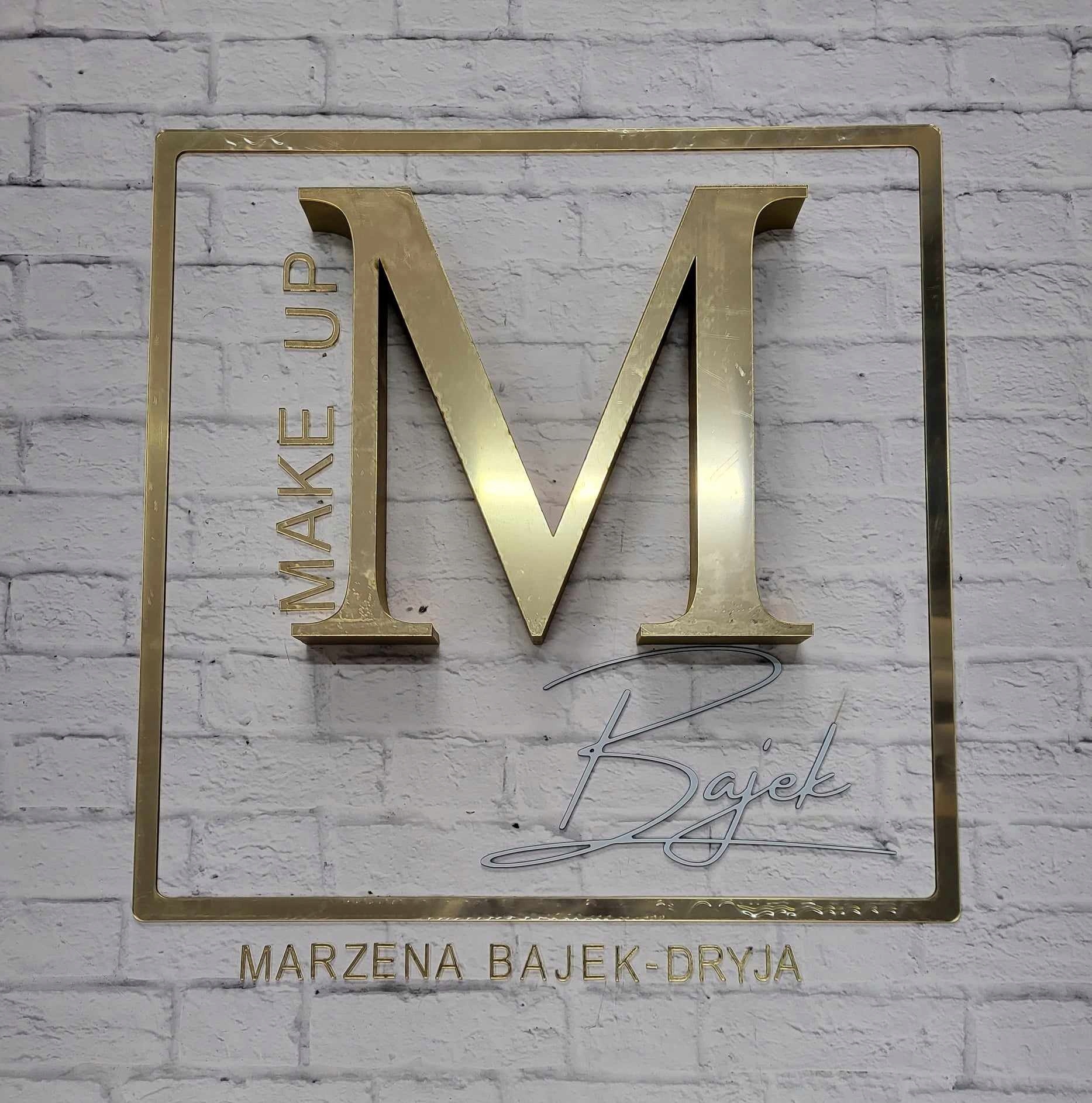 Złoty szyld z literą M, napisem MAKE UP Bajek oraz imieniem i nazwiskiem Marzena Bajek-Dryja na tle białej ściany z cegły.