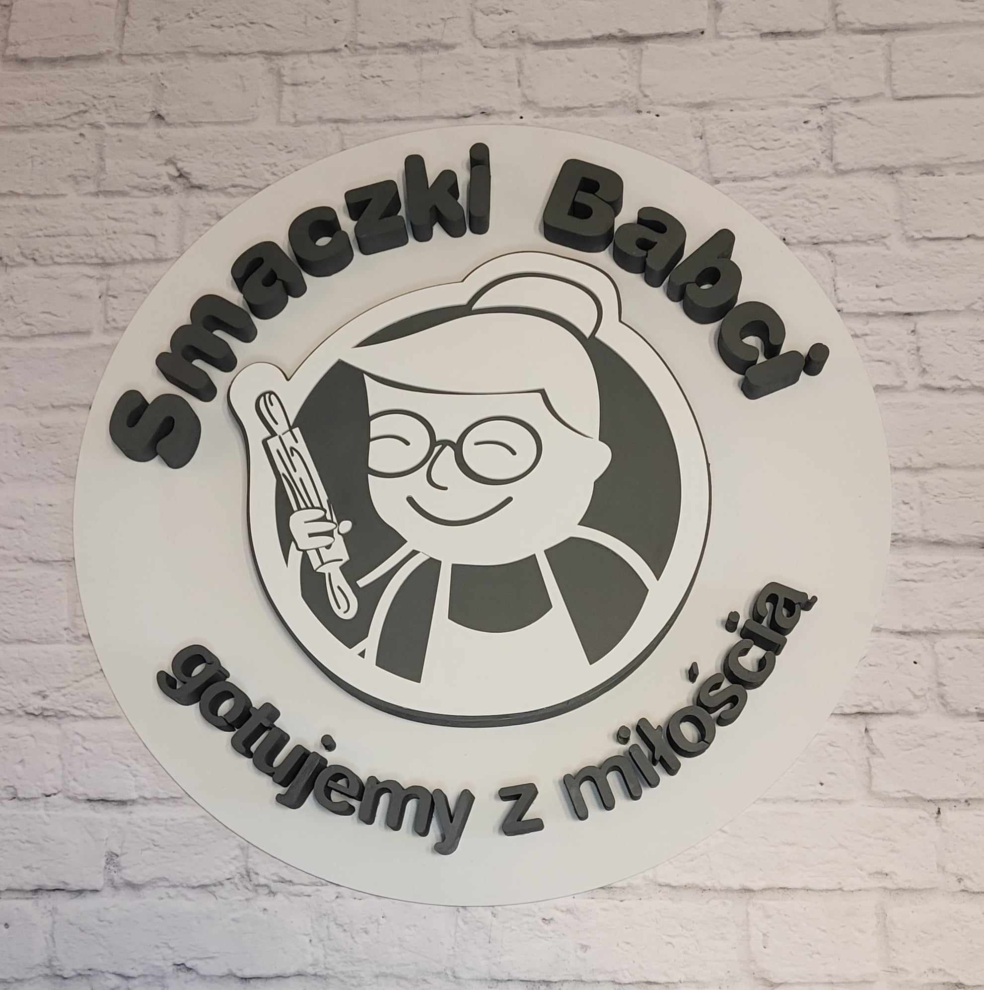 Okrągły szyld z wizerunkiem uśmiechniętej babci z wałkiem, otoczony napisem 'Smaczki Babci gotujemy z miłością', umieszczony na ścianie z białej cegły.