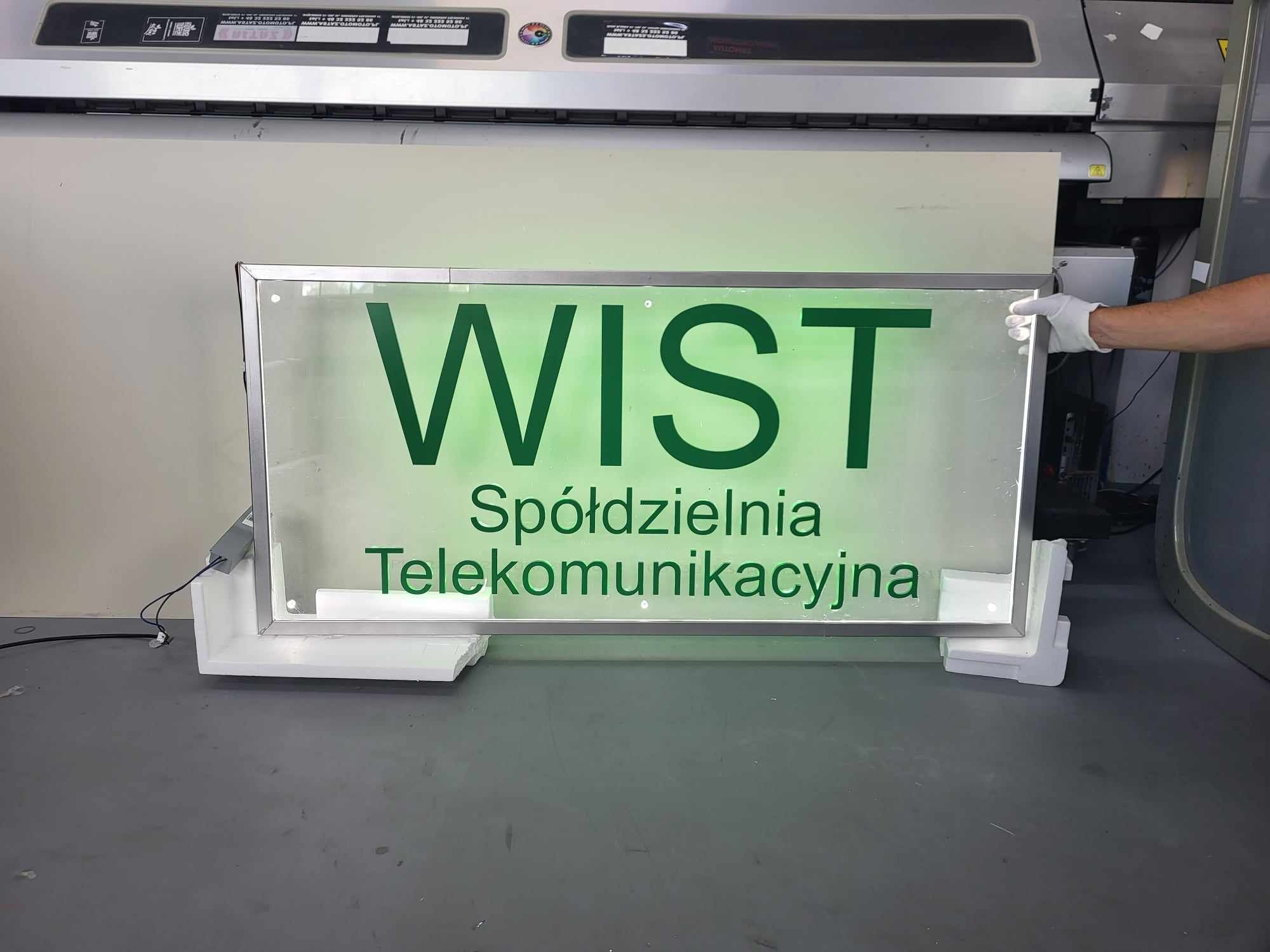 Podświetlany szyld z napisem 'WIST Spółdzielnia Telekomunikacyjna' w odcieniach zieleni, umieszczony na białych wspornikach, widoczna ręka w białej rękawiczce przytrzymująca ramę, tło stanowi...