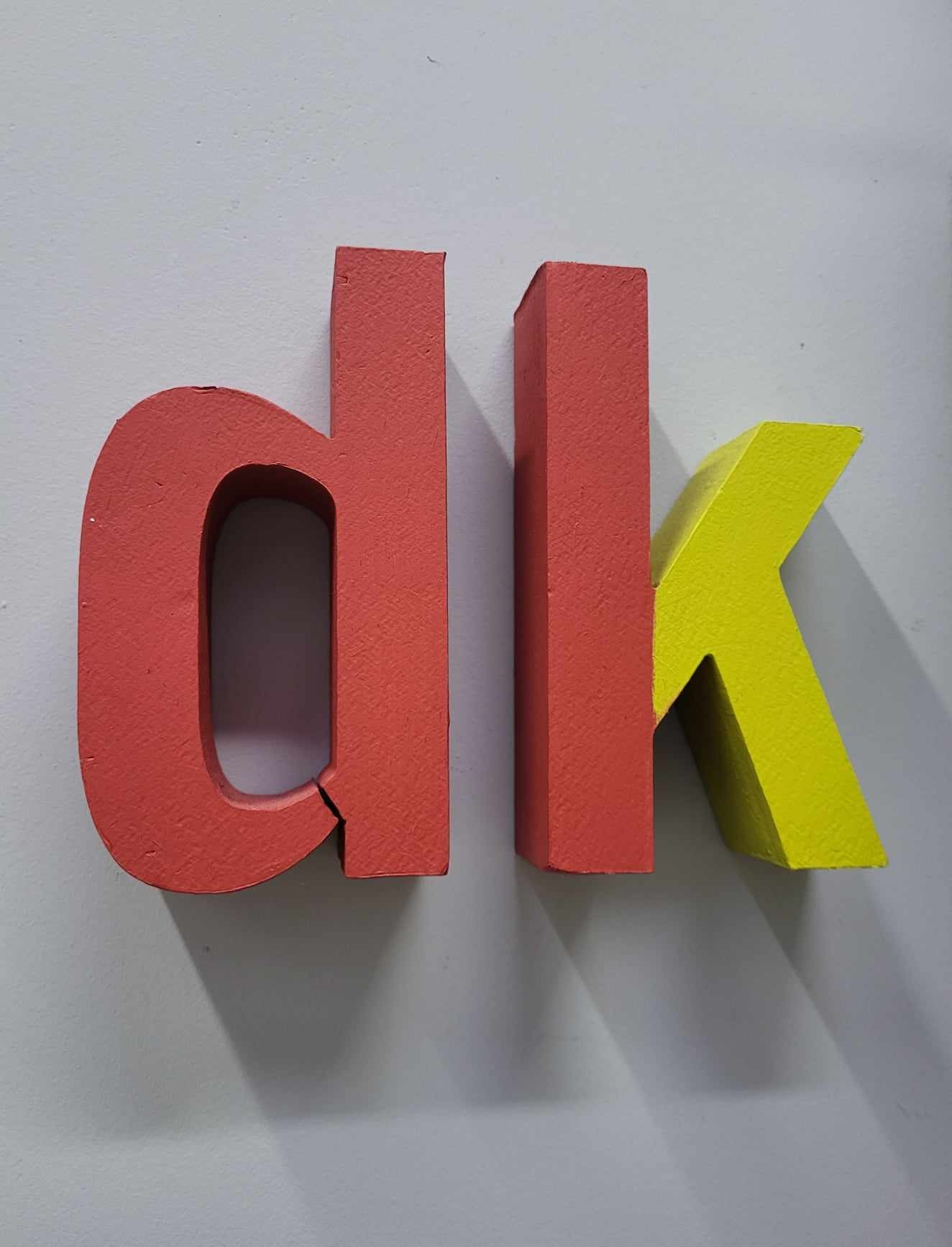 Trójwymiarowe litery 'dk' w kolorach czerwonym i żółtym, wykonane z malowanego drewna, ustawione na szarym tle, z widocznym pęknięciem na literze 'd'.