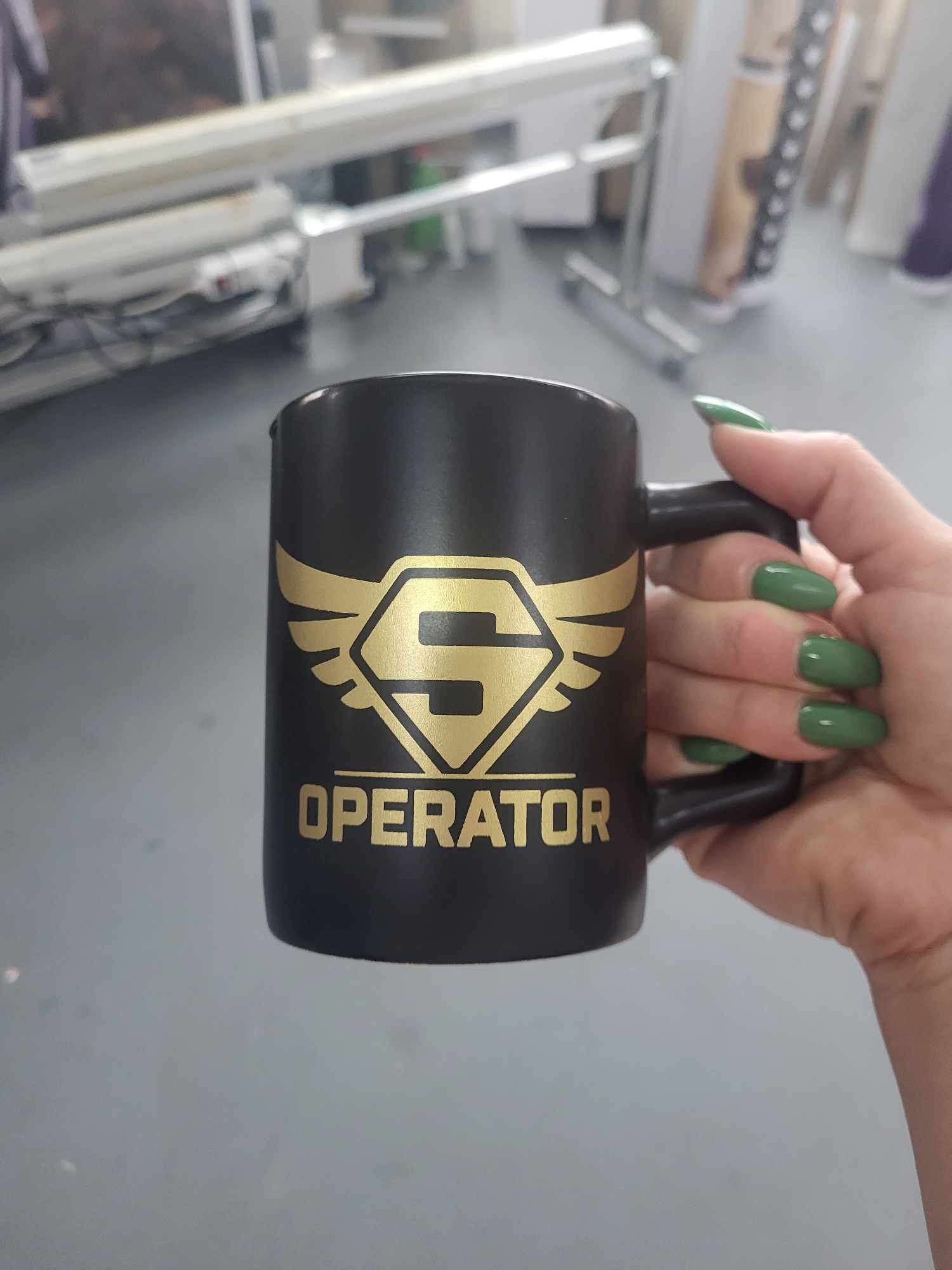 Czarny kubek z nadrukiem złotego logo i napisu 'OPERATOR' trzymany w dłoni z zielonymi paznokciami, w tle maszyny drukarskie.