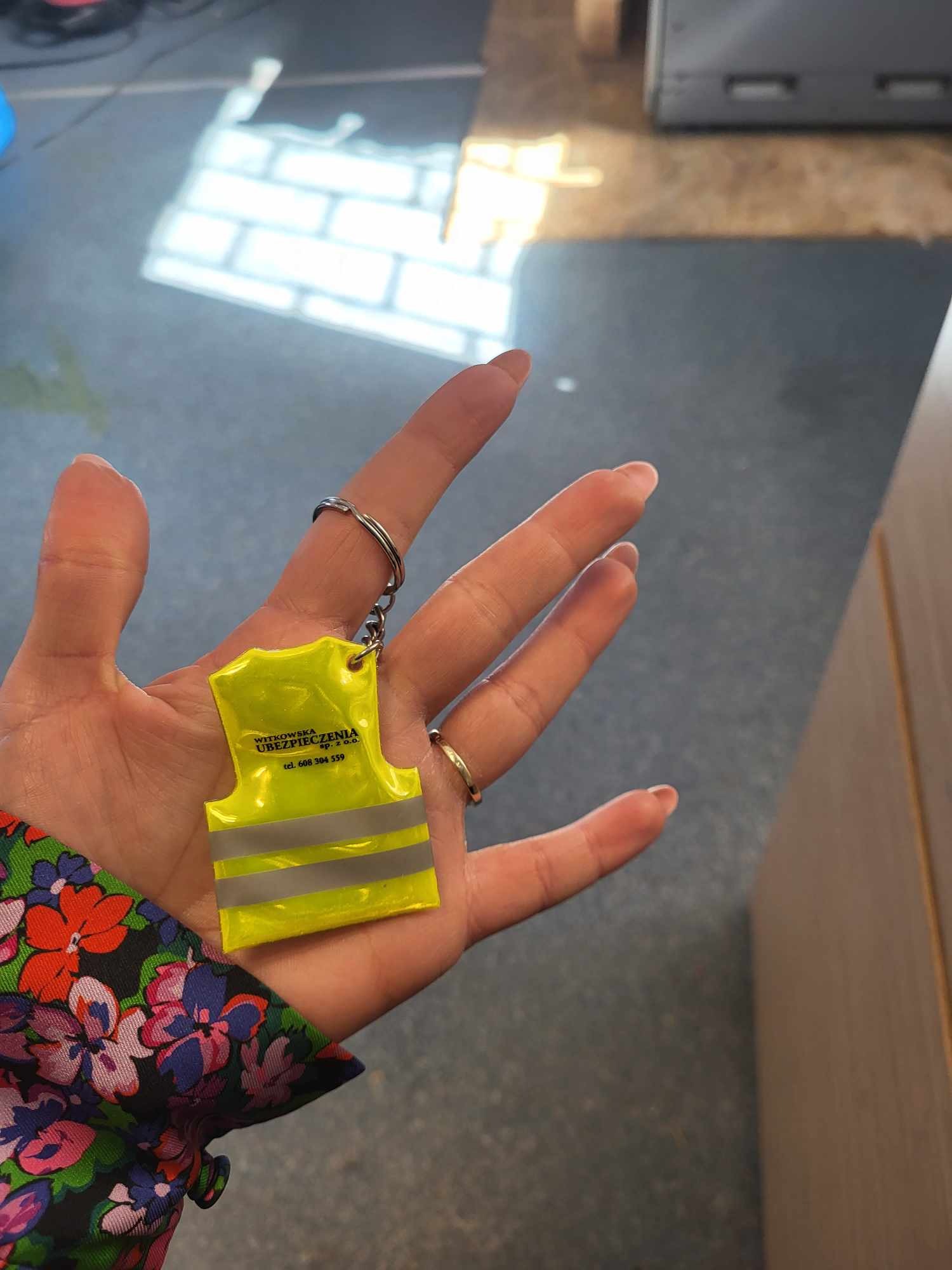 Kobieta trzyma w dłoni breloczek w kształcie miniaturowej, żółtej kamizelki odblaskowej z nadrukowanym logo firmy ubezpieczeniowej Witkowska. Breloczek zawieszony na srebrnym kółku i łańcuszku.