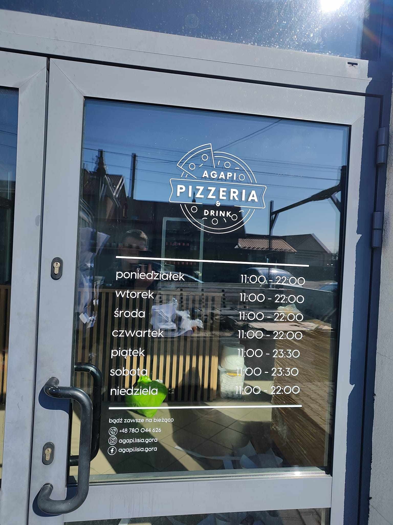 Szklane drzwi wejściowe do pizzerii z naklejonym logo i godzinami otwarcia na szybie, widoczne odbicia otoczenia.