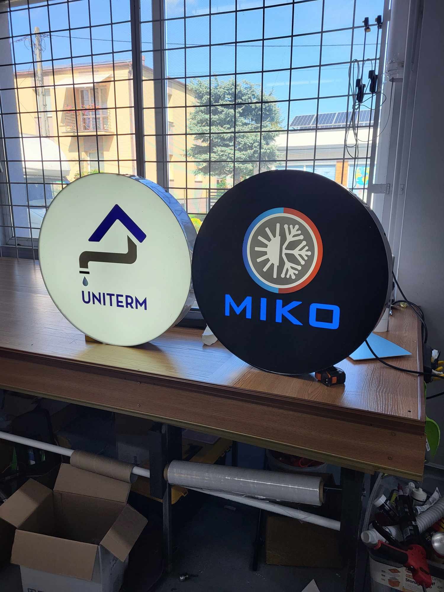 Dwa okrągłe szyldy reklamowe z podświetlanymi logotypami firm Uniterm i Miko, ustawione na stole w warsztacie, z widokiem na budynek i drzewo za oknem.