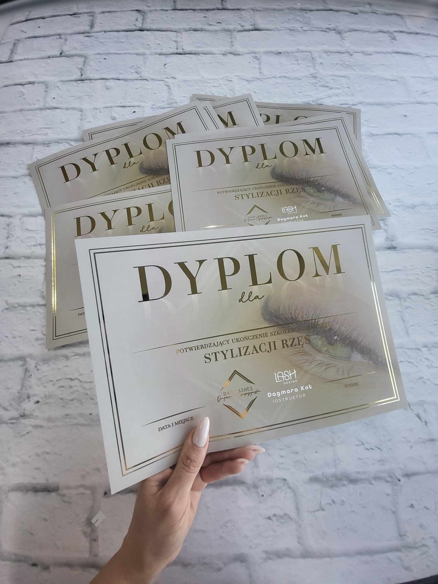 Stos dyplomów ukończenia szkolenia stylizacji rzęs, trzymanych w dłoni na tle ściany z białej cegły