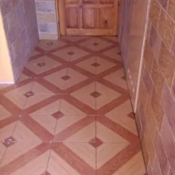 Wykończone wnętrze korytarza z brązowo-beżowymi płytkami na podłodze ułożonymi w romby oraz ścianami wyłożonymi płytkami imitującymi kamień w odcieniach brązu i beżu.