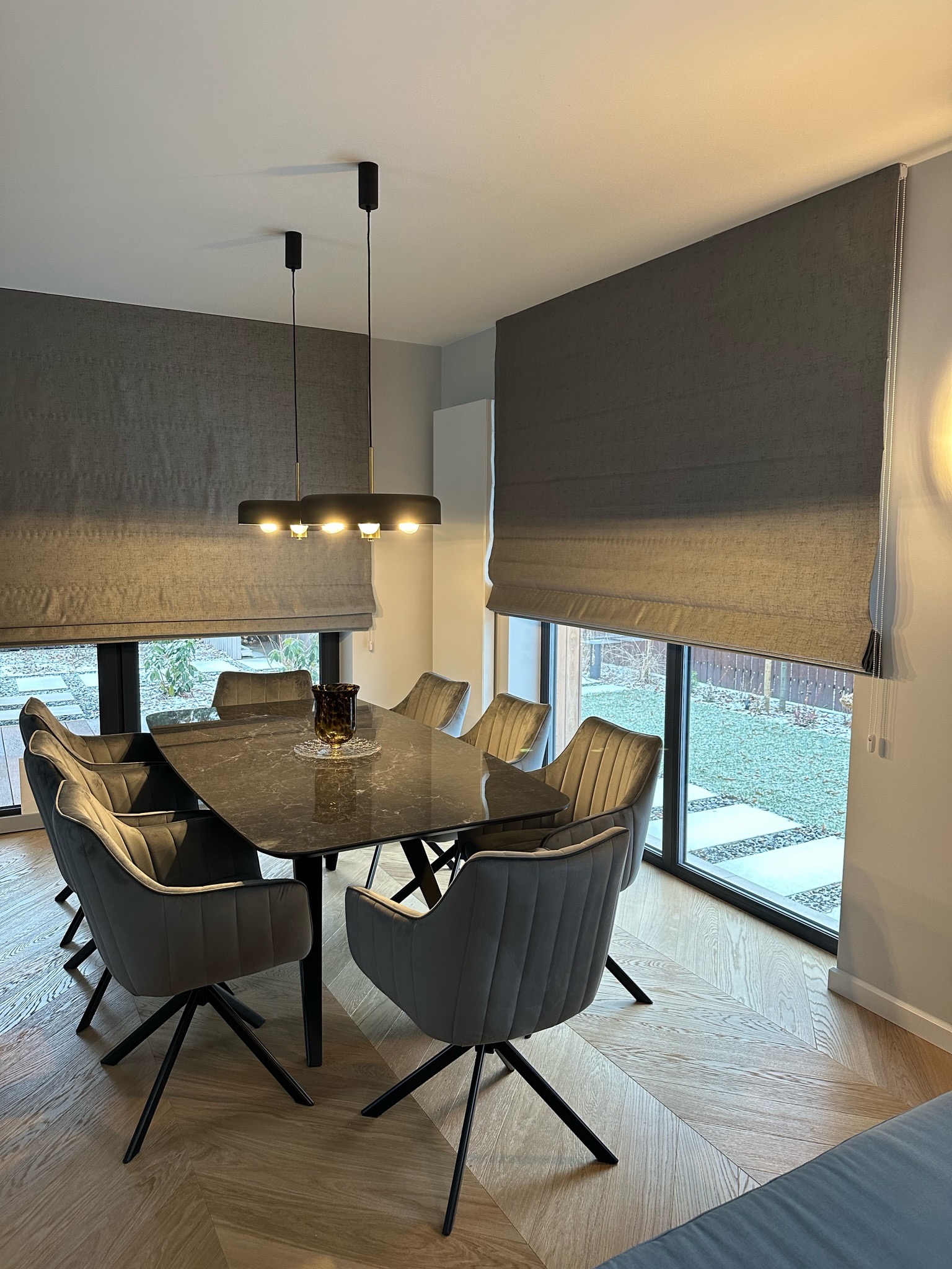 Eleganckie rolety rzymskie w kolorze szarym, zamontowane w nowoczesnym wnętrzu jadalni z dużym oknem i parkietem w jodełkę. Minimalistyczny design i stonowana kolorystyka.