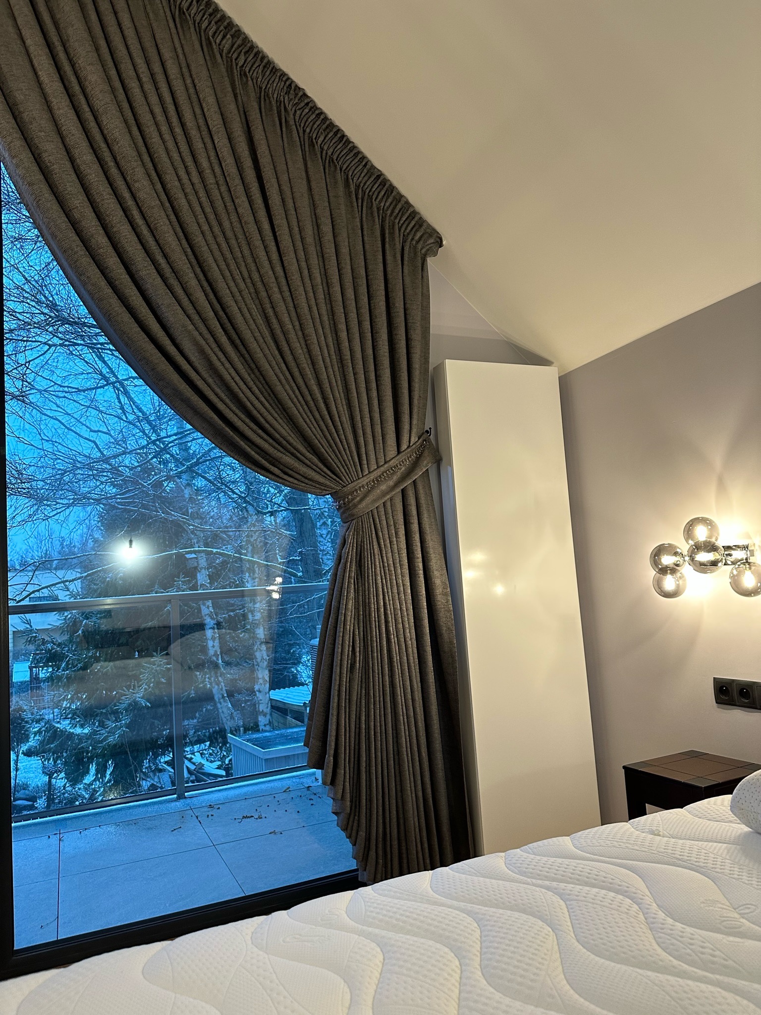 Elegancka, grafitowa zasłona upięta dekoracyjnym sznurem, odsłaniająca zimowy krajobraz za oknem balkonowym. Widok z wnętrza sypialni, z fragmentem łóżka i lampy.