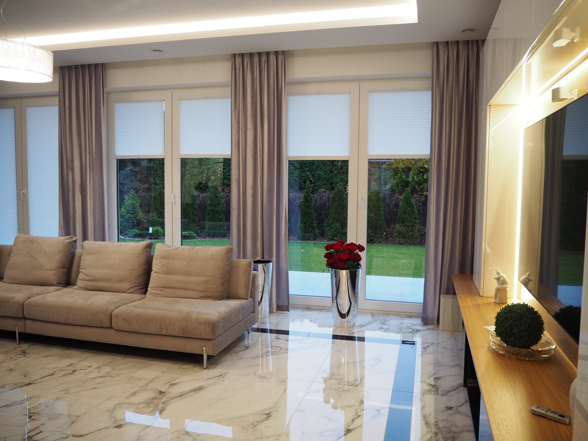 Elegancki salon z beżową sofą, marmurową podłogą i dużymi oknami z plisowanymi roletami i zasłonami w odcieniach szarości, uzupełniony srebrnymi wazonami i elementami dekoracyjnymi.