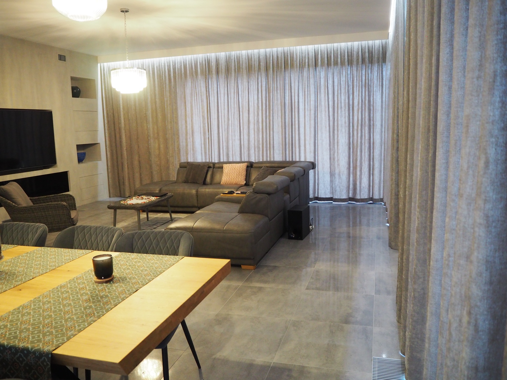 Jasne, przestronne wnętrze z beżowymi zasłonami na całej ścianie, szarym narożnikiem i drewnianym stołem z dekoracyjnym bieżnikiem.