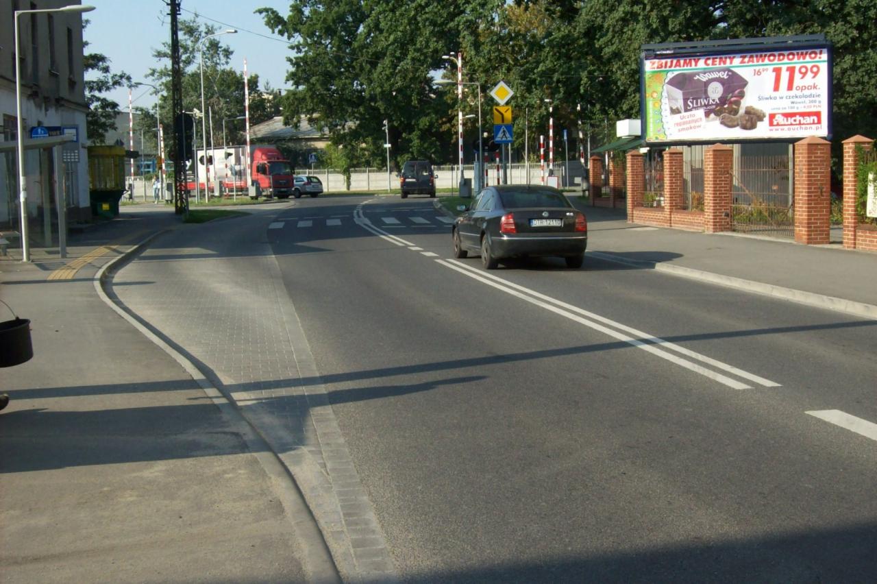 Ulica z przejściem dla pieszych i samochodem osobowym w tle, obok billboard reklamowy słodyczy Auchan.