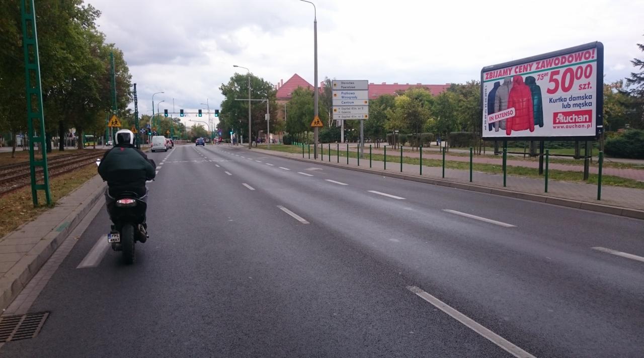 Ulica miejska z billboardem reklamowym Auchan, na pierwszym planie motocyklista w kasku, w tle budynek z czerwonym dachem i znaki drogowe.