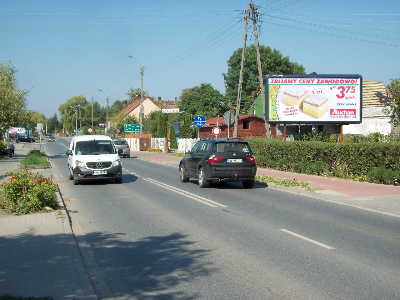 Billboard reklamujący kremówki z Auchan, umieszczony przy drodze w wiejskiej scenerii, widoczny z perspektywy ulicy, z przejeżdżającymi samochodami.