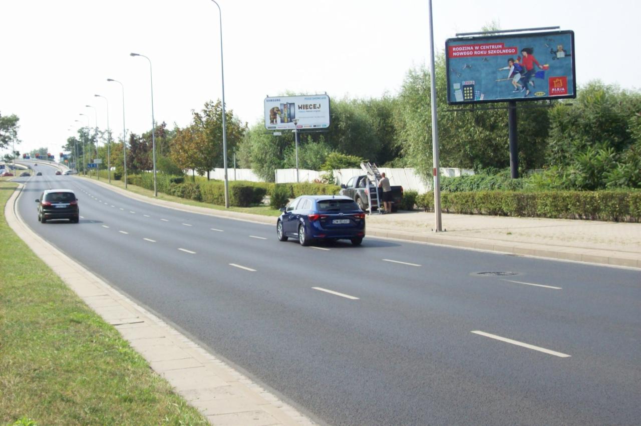 Ulica miejska z dwoma bilbordami reklamowymi w tle, pracownik na drabinie przy jednym z nich, niebieski samochód jedzie w kierunku widza.