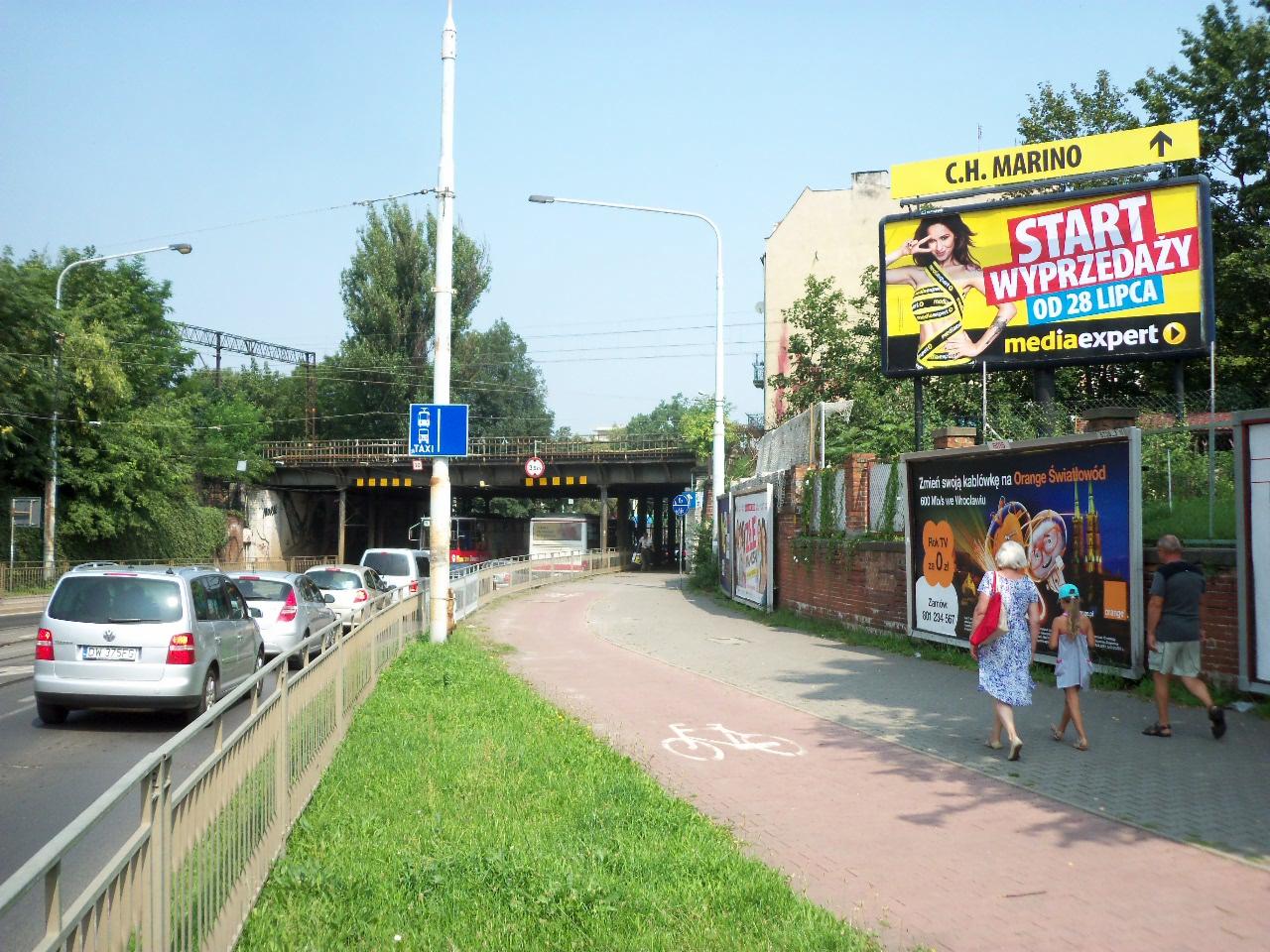 Billboard z reklamą wyprzedaży Media Expert z modelką w żółtej sukience, widoczny z ulicy z przejeżdżającymi samochodami i przechodniami na chodniku.