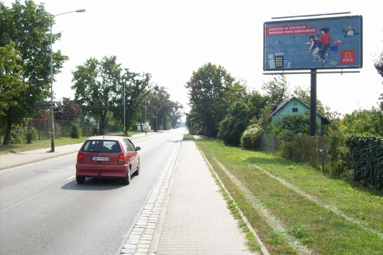 Billboard z reklamą 'Rodzina w centrum nowego roku szkolnego' umieszczony przy drodze, obok przejeżdża czerwony samochód, w tle zieleń i budynek.