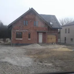 Surowy stan dwukondygnacyjnego domu z czerwonej cegły z zamontowanymi oknami i pokrytym dachem z dachówką, garaż z drewnianymi wrotami, widoczne pozostałości materiałów budowlanych na posesji.