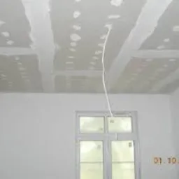 Sufit z płyt kartonowo-gipsowych w trakcie szpachlowania, widoczne łączenia płyt, kabel wystający z sufitu, białe ściany i okno w tle.