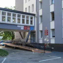 Zewnętrzny łącznik między budynkami z licznymi oknami, widoczny numer budynku 13 i podjazd dla niepełnosprawnych.