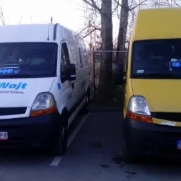 Dwa busy Renault Master zaparkowane obok siebie: biały z niebieskim logo firmy transportowej 'Big-Wojt' i żółty z podświetlanym napisem 'Big Wojt' na desce rozdzielczej. W tle widoczne drzewa...