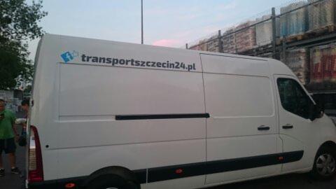 Biały bus dostawczy z niebieskim logo i napisem transportszczecin24.pl, zaparkowany na tle magazynu z paletami, widoczne osoby w tle.