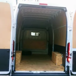 Wnętrze pustego, wyłożonego sklejką białego busa z otwartymi tylnymi drzwiami. Na podłodze widoczne dwie drewniane skrzynie i opona.