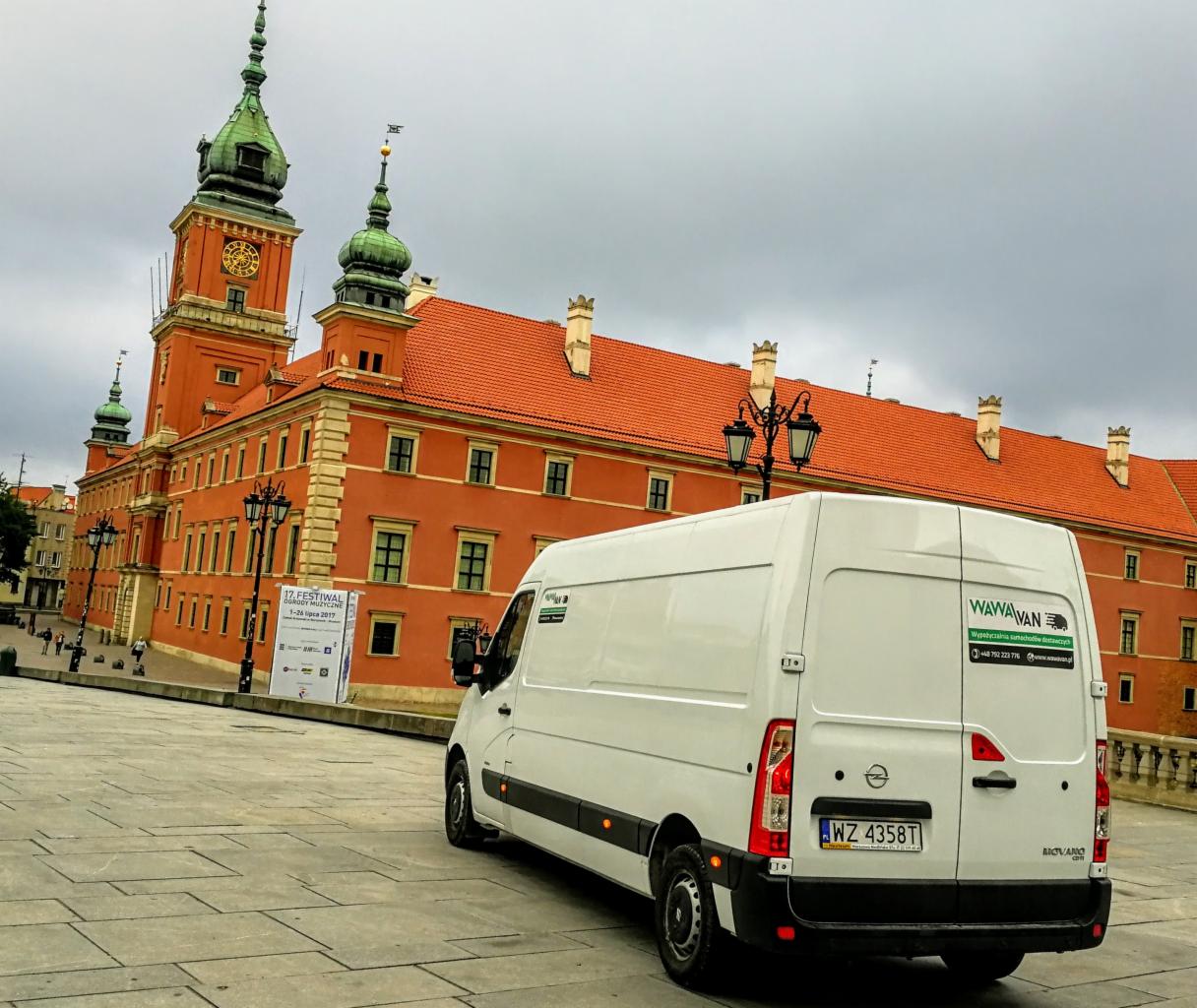 Biały samochód dostawczy z logo firmy na tle Zamku Królewskiego w Warszawie, pochmurne niebo.