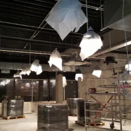 Wnętrze komercyjne w trakcie instalacji oświetlenia, z lampami zabezpieczonymi folią, rusztowaniem i materiałami budowlanymi