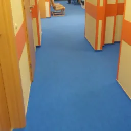 Korytarz w budynku w Warszawie z nową, intensywnie niebieską podłogą tekstylną, widoczne drzwi i pomarańczowe akcenty na ścianach.