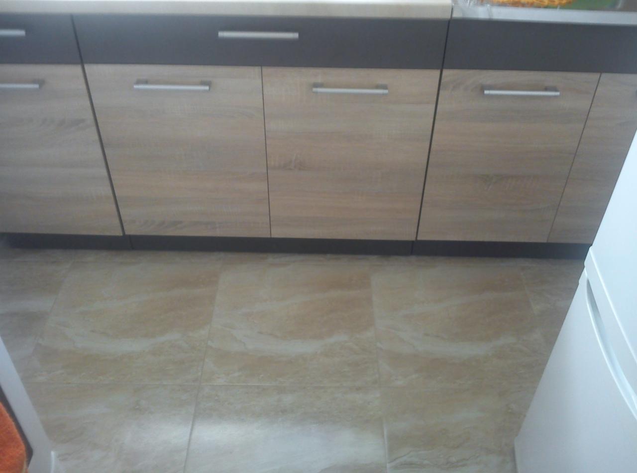 Częściowe ujęcie dolnych szafek kuchennych z jasnego drewna z szarymi górnymi szufladami i beżowej podłogi z płytek imitujących marmur.