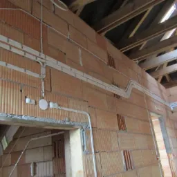 Wnętrze w trakcie budowy z widocznymi przewodami elektrycznymi poprowadzonymi po ścianach z cegły dziurawki, z widocznymi puszkami instalacyjnymi i elementami drewnianej konstrukcji dachu.