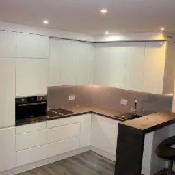 Nowoczesna, jasna kuchnia z białymi, lakierowanymi frontami, szarym blatem i barkiem z dwoma hokerami. Oświetlenie punktowe podkreśla minimalistyczny design.