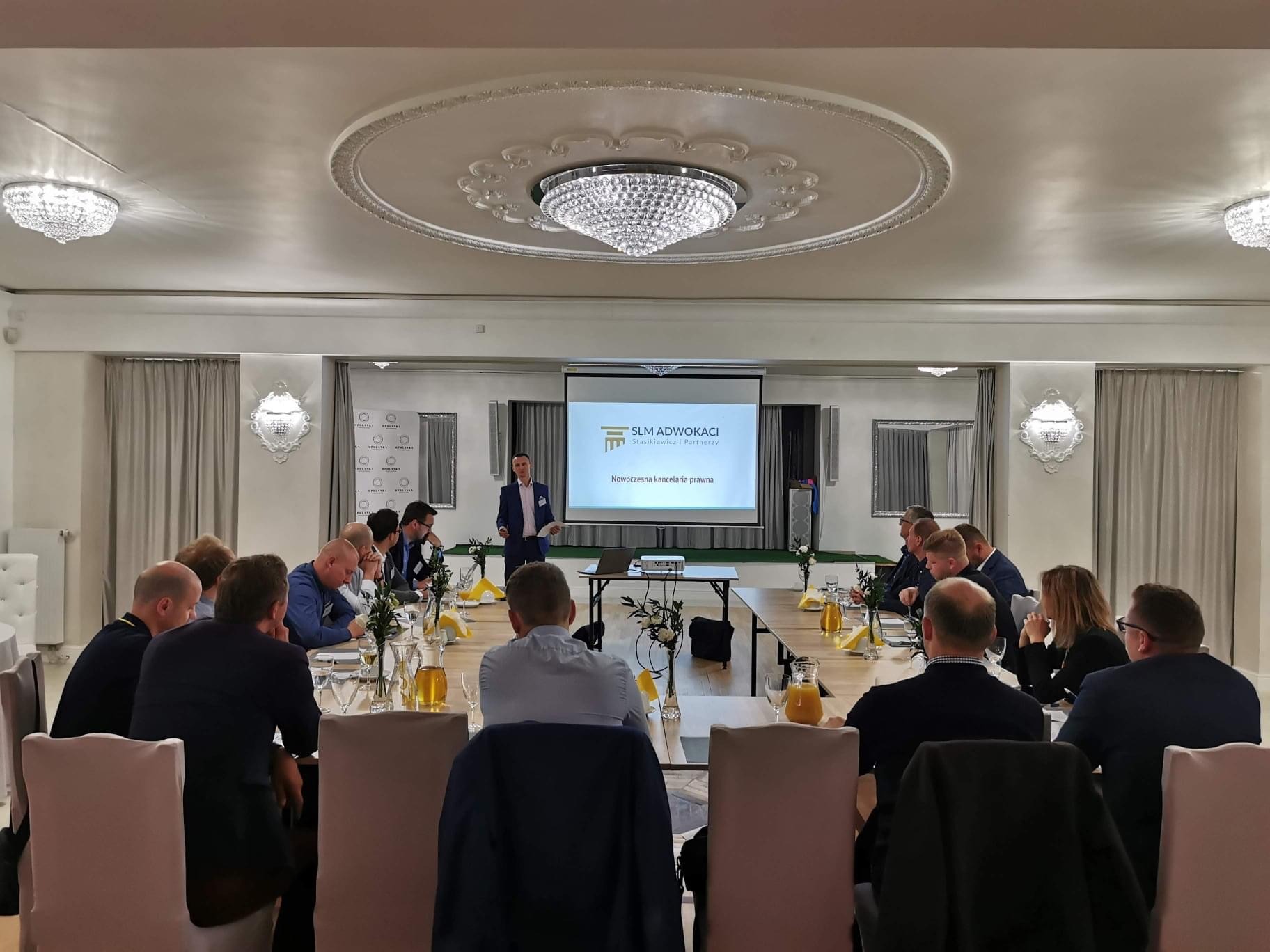 Prezentacja kancelarii prawnej SLM Adwokaci podczas spotkania biznesowego, uczestnicy siedzą przy stole z napojami, na ekranie wyświetlone logo i hasło 'Nowoczesna kancelaria prawna'.