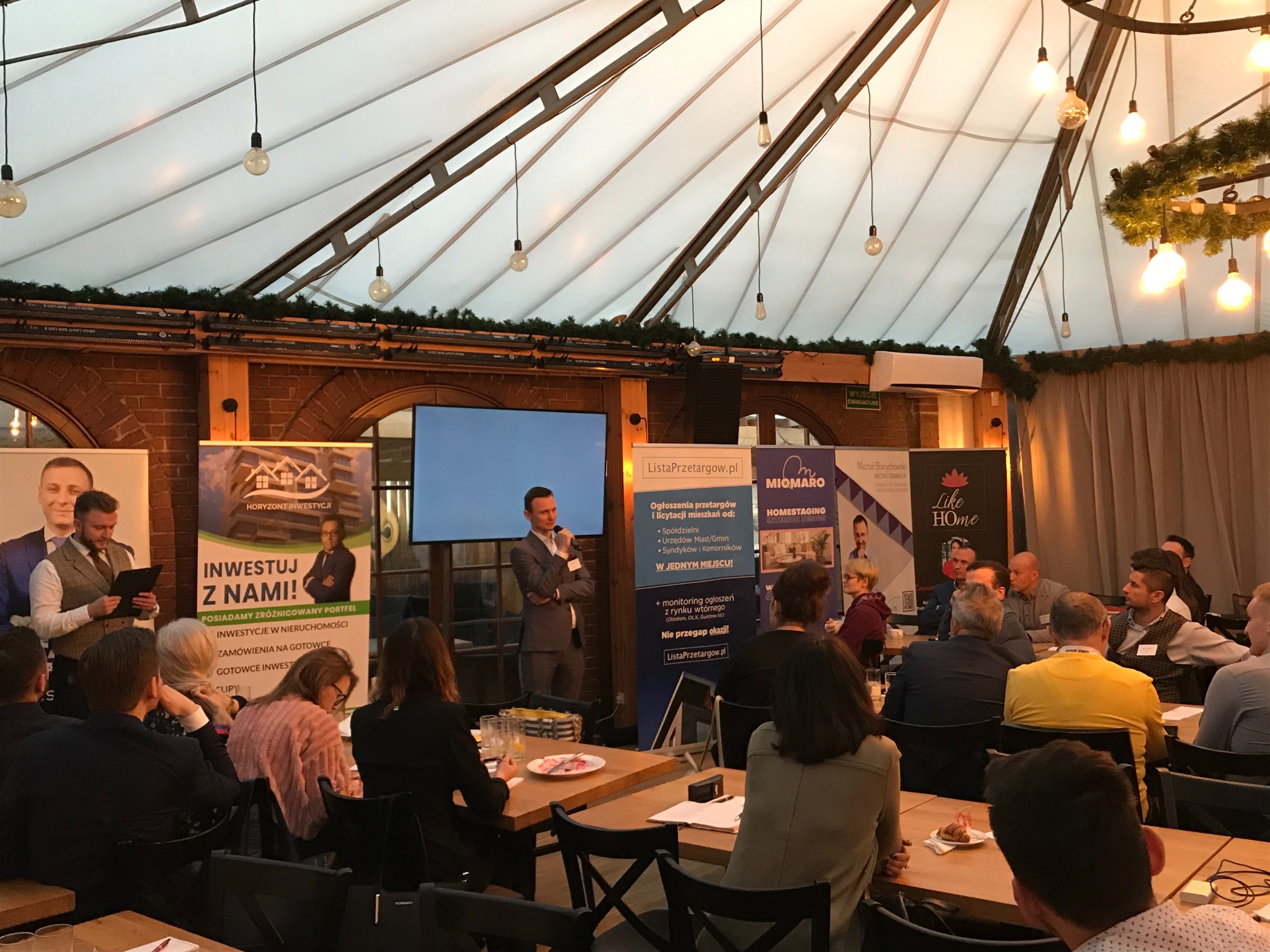 Konferencja o inwestycjach w nieruchomości w Opolu, prelegent w szarym garniturze przemawia do publiczności, w tle banery reklamowe i dekoracyjne oświetlenie.