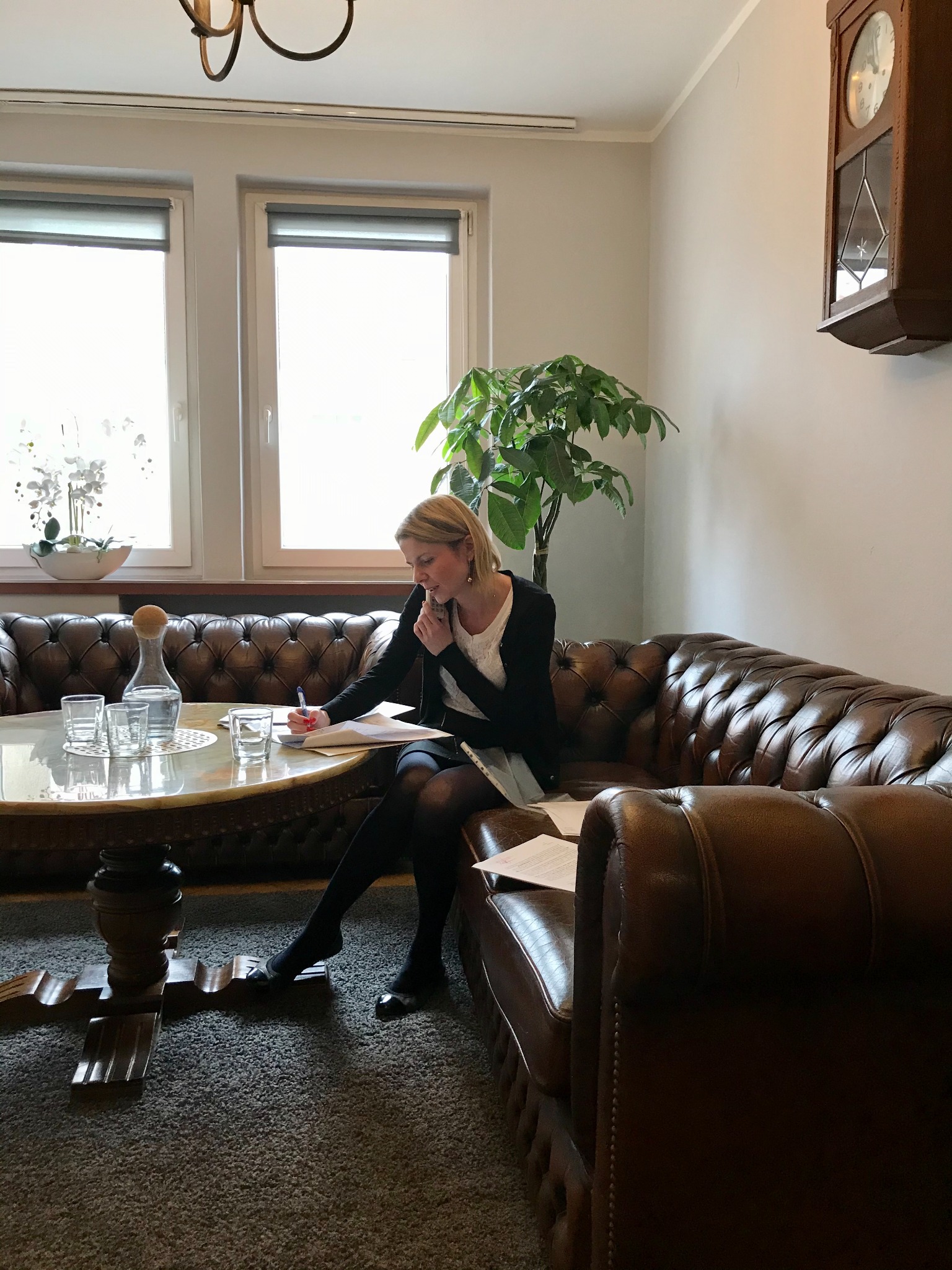 Kobieta w biurze, siedząca na skórzanej sofie, analizuje dokumenty przy okrągłym stoliku z szklankami wody, w tle widoczny zegar i roślina doniczkowa.